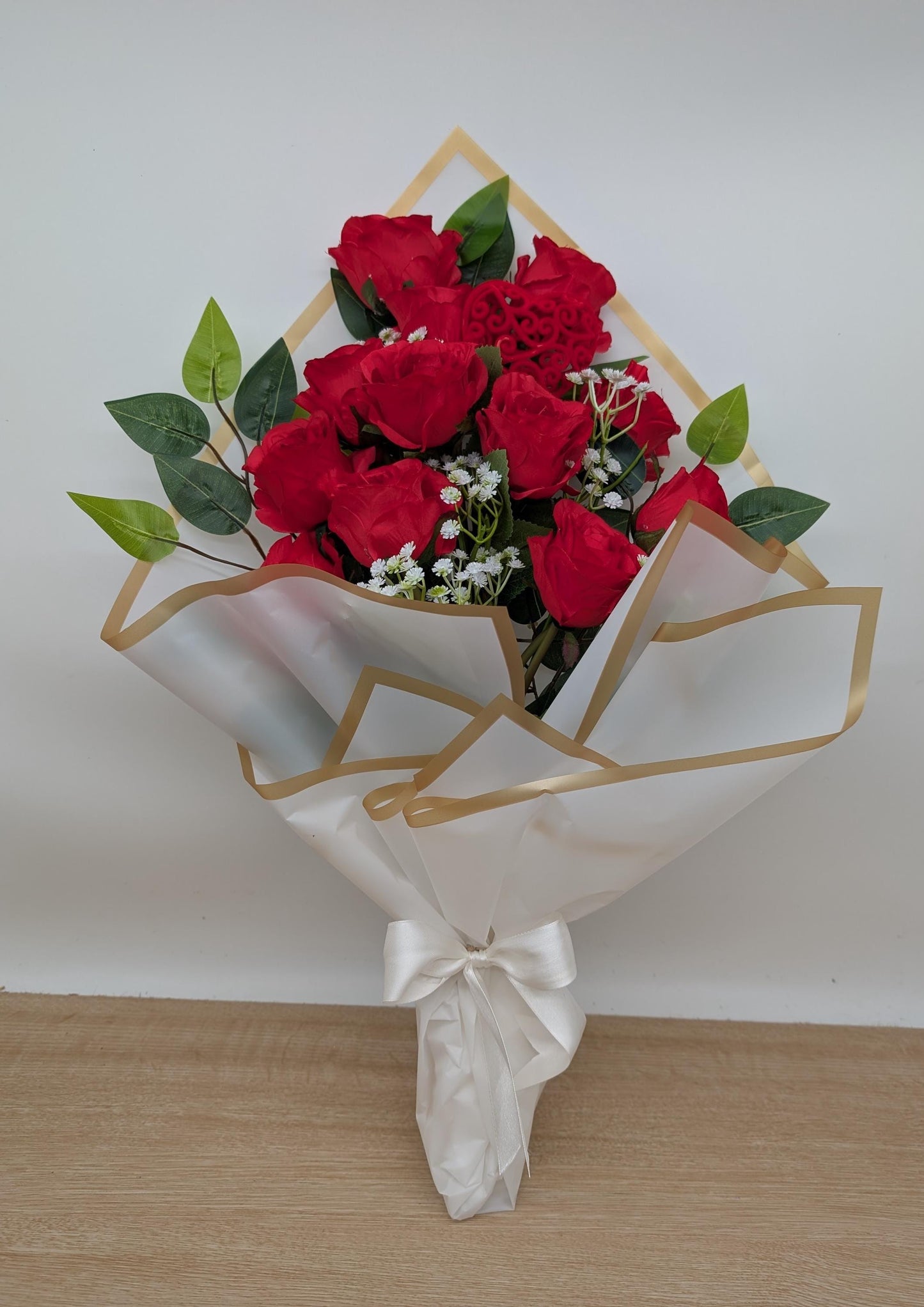 Bouquet of red roses with green leaves wrapped in white paper with gold geometric patterns on a light wooden floor.