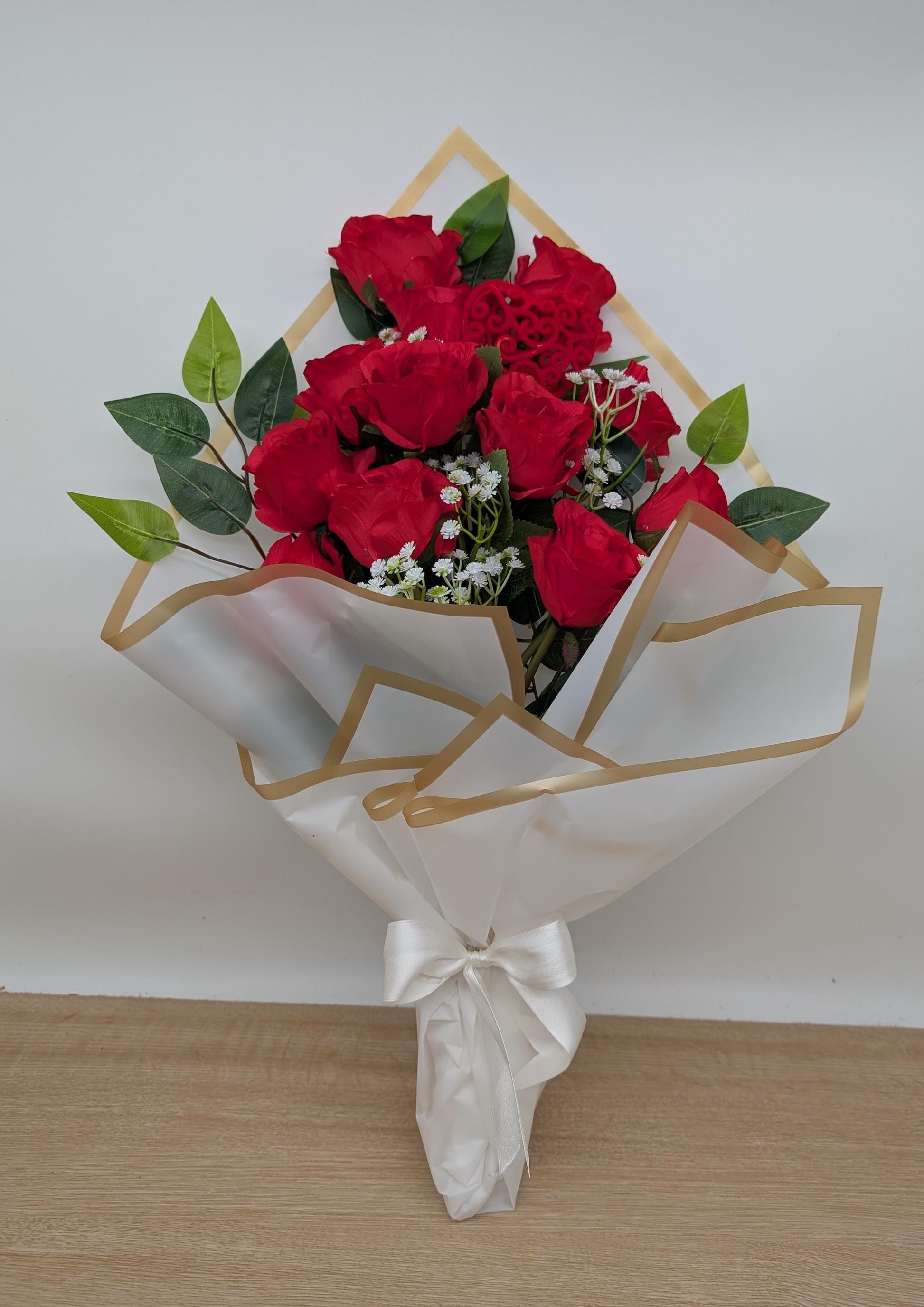 Bouquet of red roses with green leaves wrapped in white paper with gold geometric patterns on a light wooden floor.