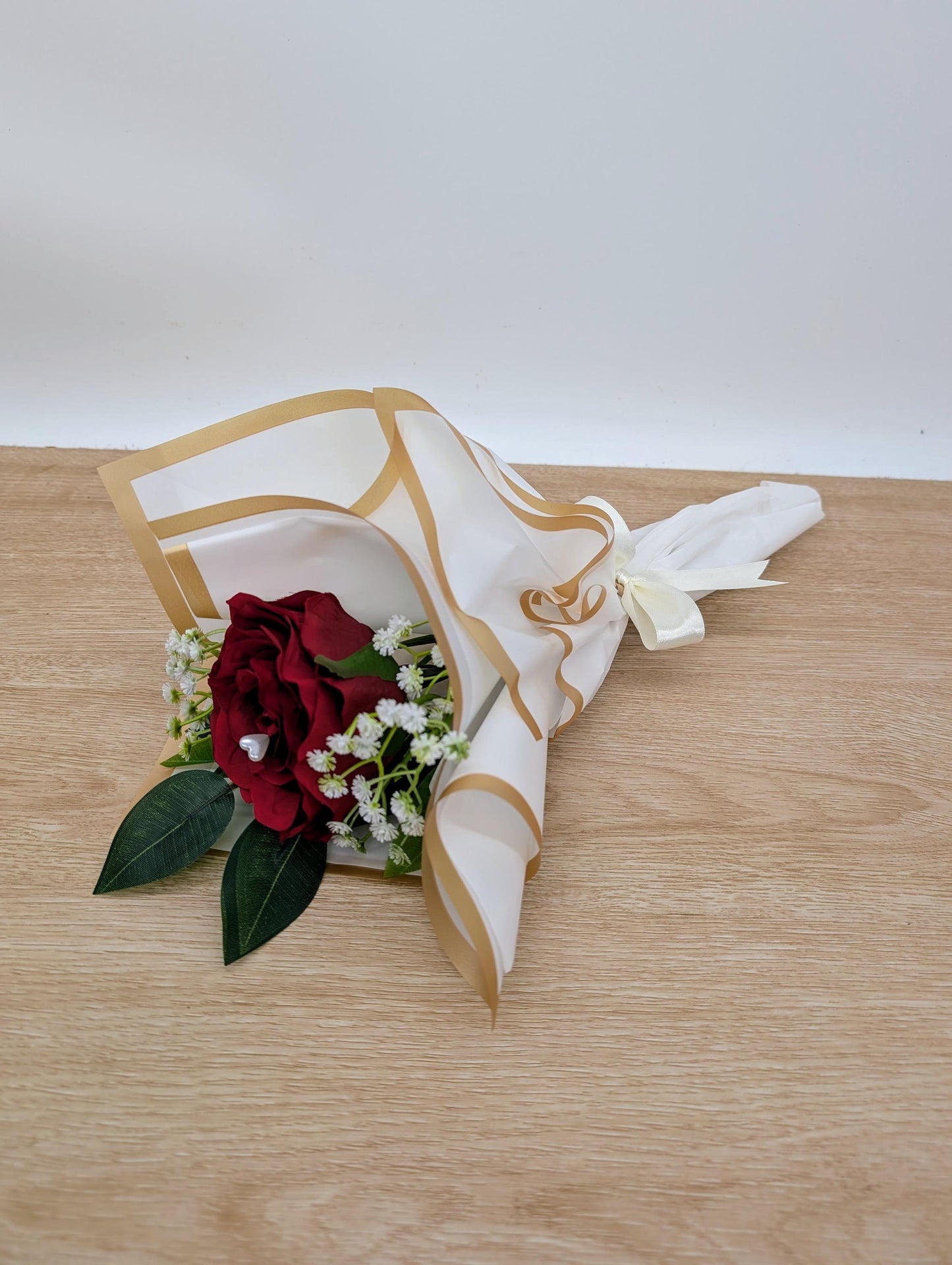 Floral arrangement with a red rose and green leaves in a decorative white and gold container on a wooden surface.