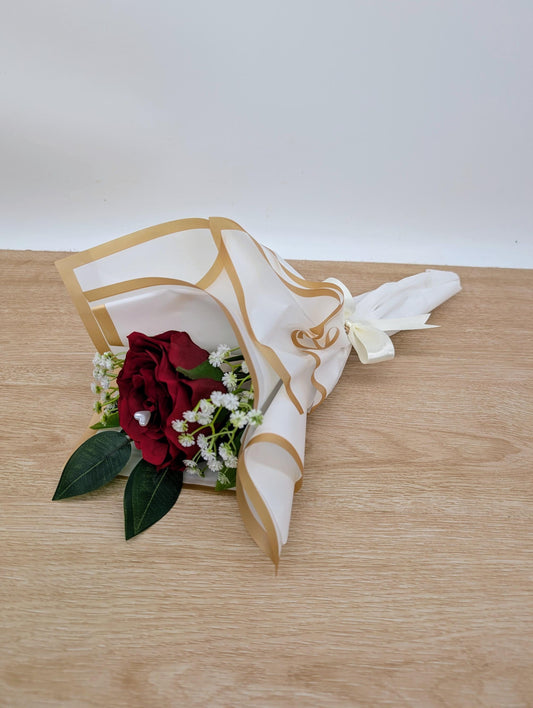 Floral arrangement with a red rose and green leaves in a decorative white and gold container on a wooden surface.