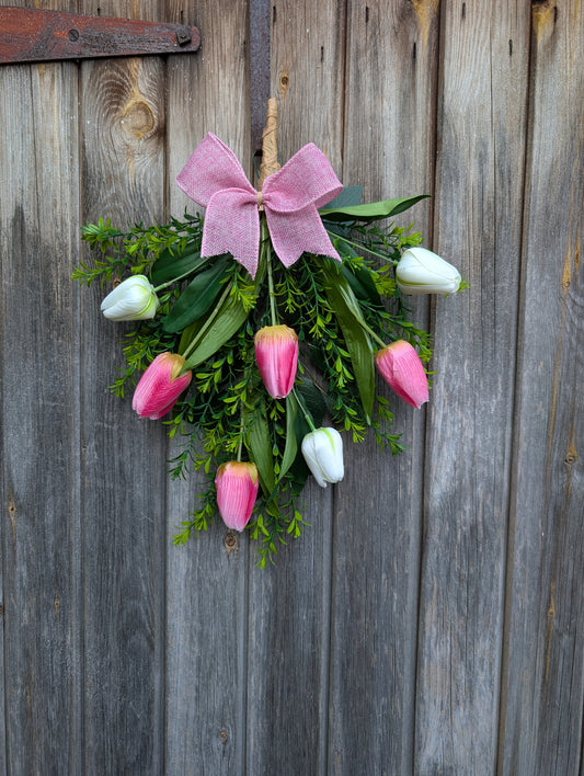 Rustic spring tulip door swag with pink and white tulips, greenery, and a pink burlap bow hanging on a wooden fence.