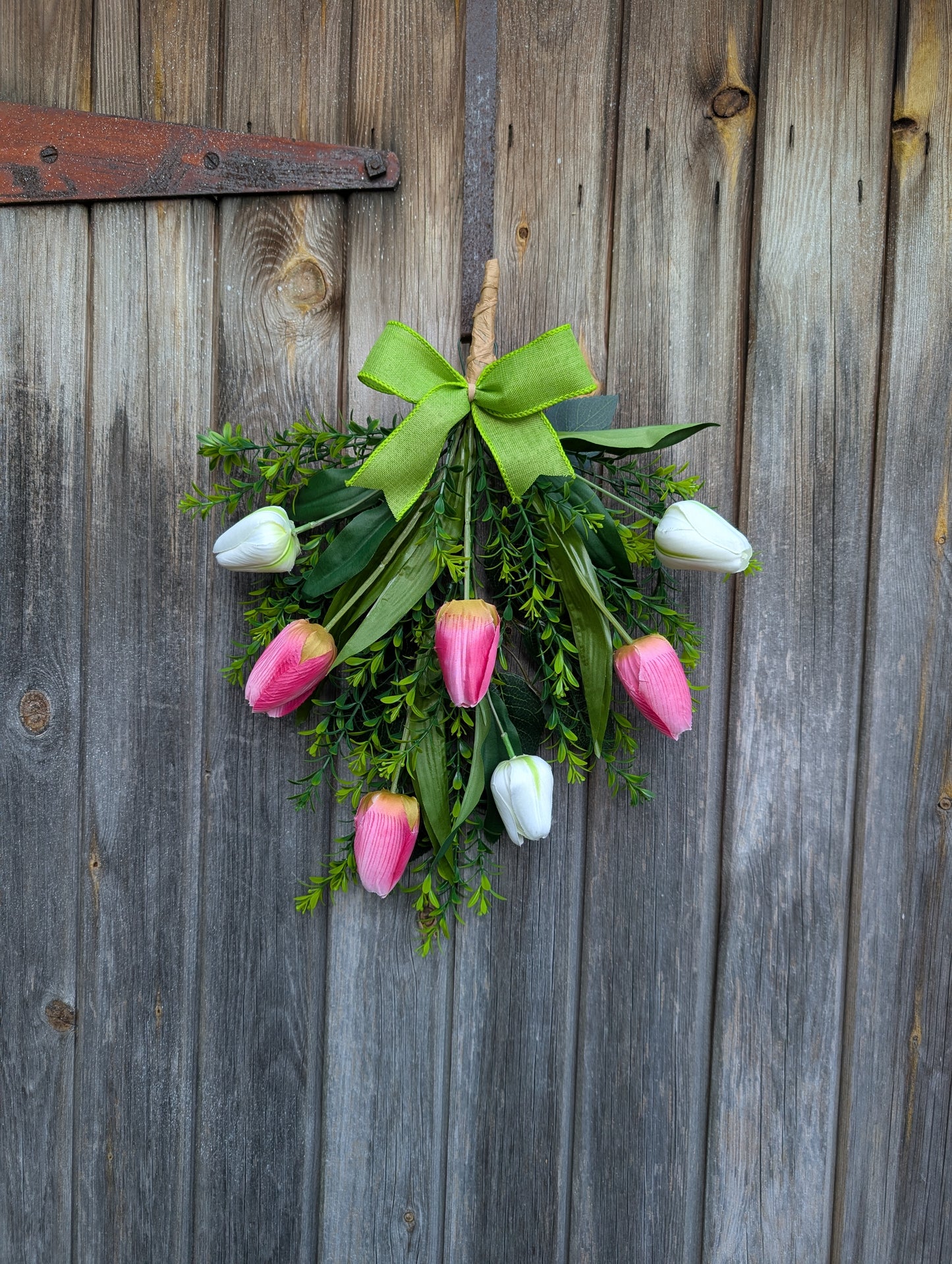 Rustic spring tulip door swag with pink and white tulips, greenery, and a pink burlap bow hanging omn a wooden door 