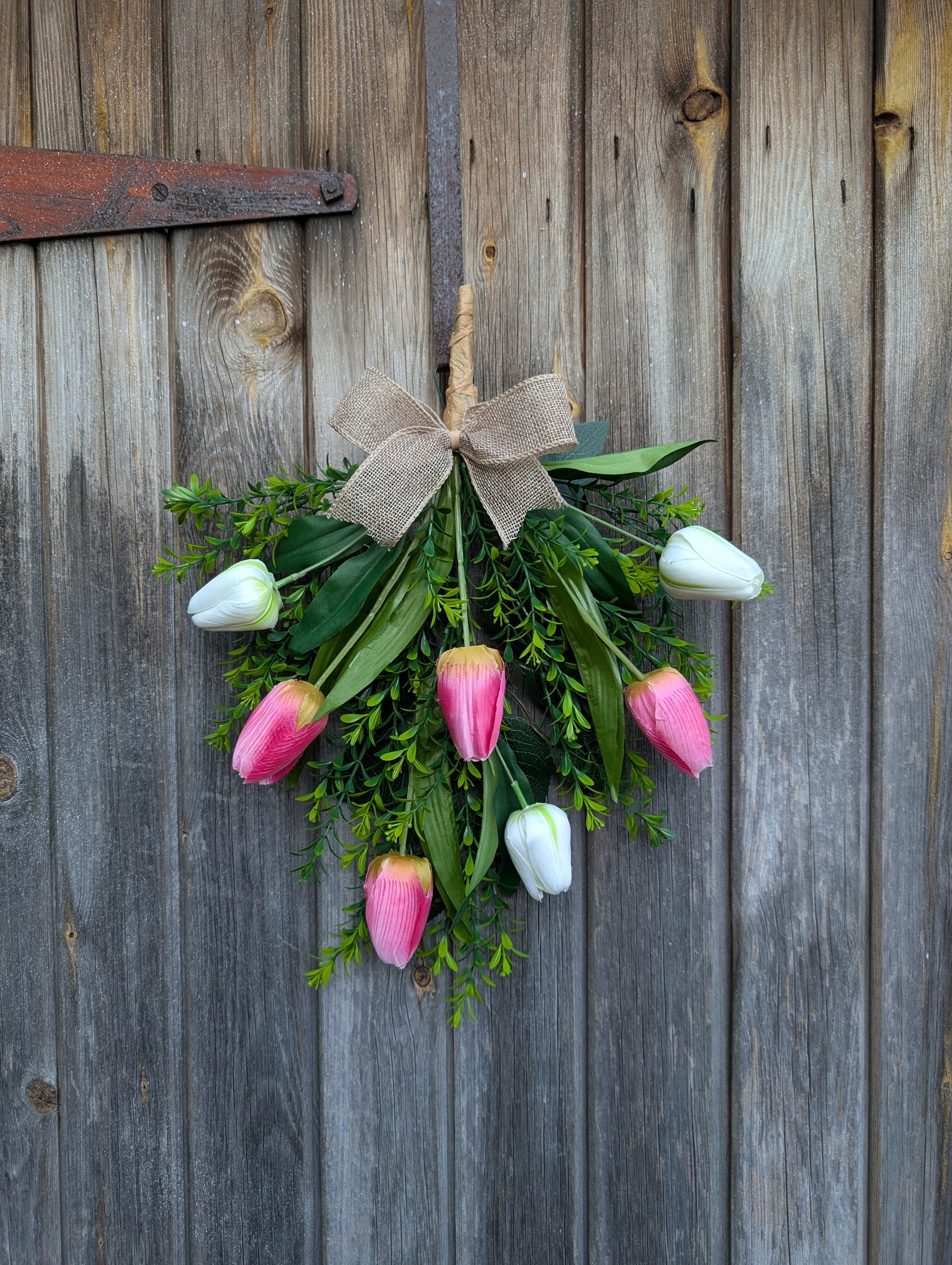 Rustic spring tulip door swag with pink and white tulips, greenery, and a pink burlap bow hanging omn a wooden door 